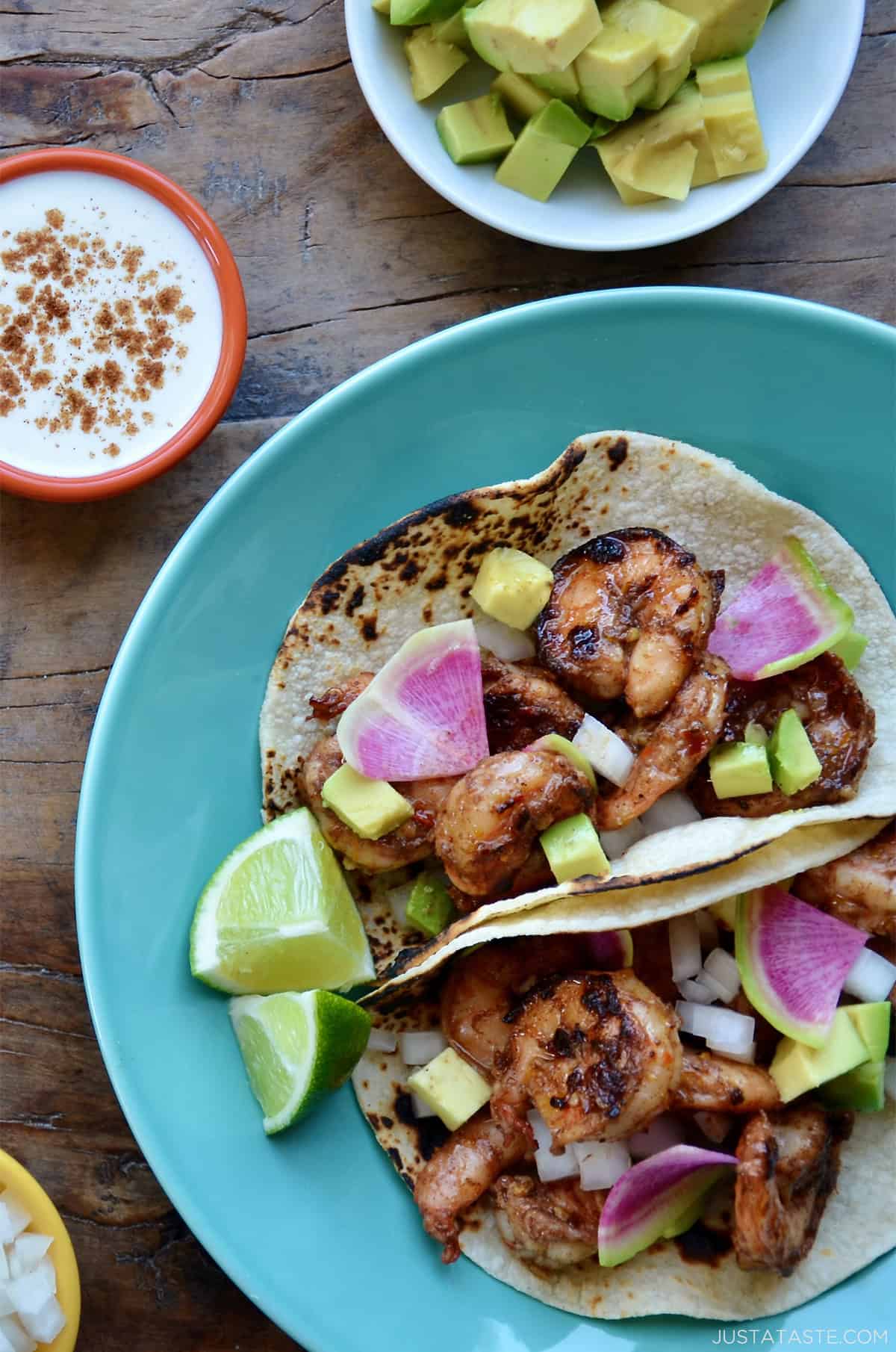 Spicy shrimp tacos in corn tortillas topped with a lime crema, sliced radishes and avocado on a dinner plate.