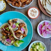 A top-down view of spicy shrimp tacos next to a plate piled high with spicy shrimp and small bowls containing diced avocado, slices of watermelon radish, and lime crema.