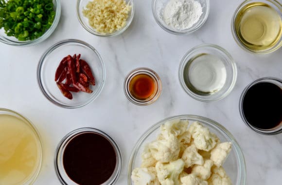 Various sized glass bowls containing chopped scallions, minced ginger, cornstarch, vegetable oil, hoisin sauce, sesame oil, rice wine vinegar, vegetable stock and cauliflower florets 