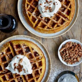 Two plates each hold a Pumpkin Spice Waffle, topped with a dollop of whipped cream, a drizzle of syrup, and a sprinkle of pecans. Nearby is a glass pitcher of maple syrup, a dish of chopped nuts, and a sifter with powdered sugar.