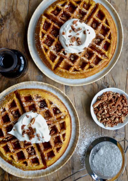 Two plates each hold a Pumpkin Spice Waffle, topped with a dollop of whipped cream, a drizzle of syrup, and a sprinkle of pecans. Nearby is a glass pitcher of maple syrup, a dish of chopped nuts, and a sifter with powdered sugar.