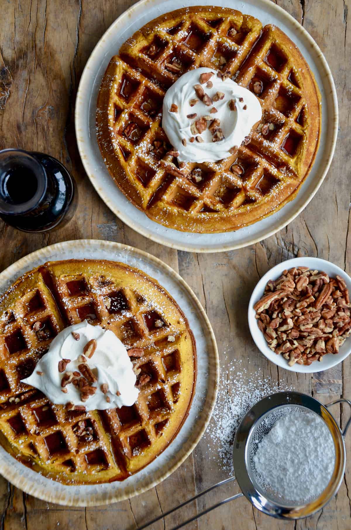 Two plates each hold a Pumpkin Spice Waffle, topped with a dollop of whipped cream, a drizzle of syrup, and a sprinkle of pecans. Nearby is a glass pitcher of maple syrup, a dish of chopped nuts, and a sifter with powdered sugar.