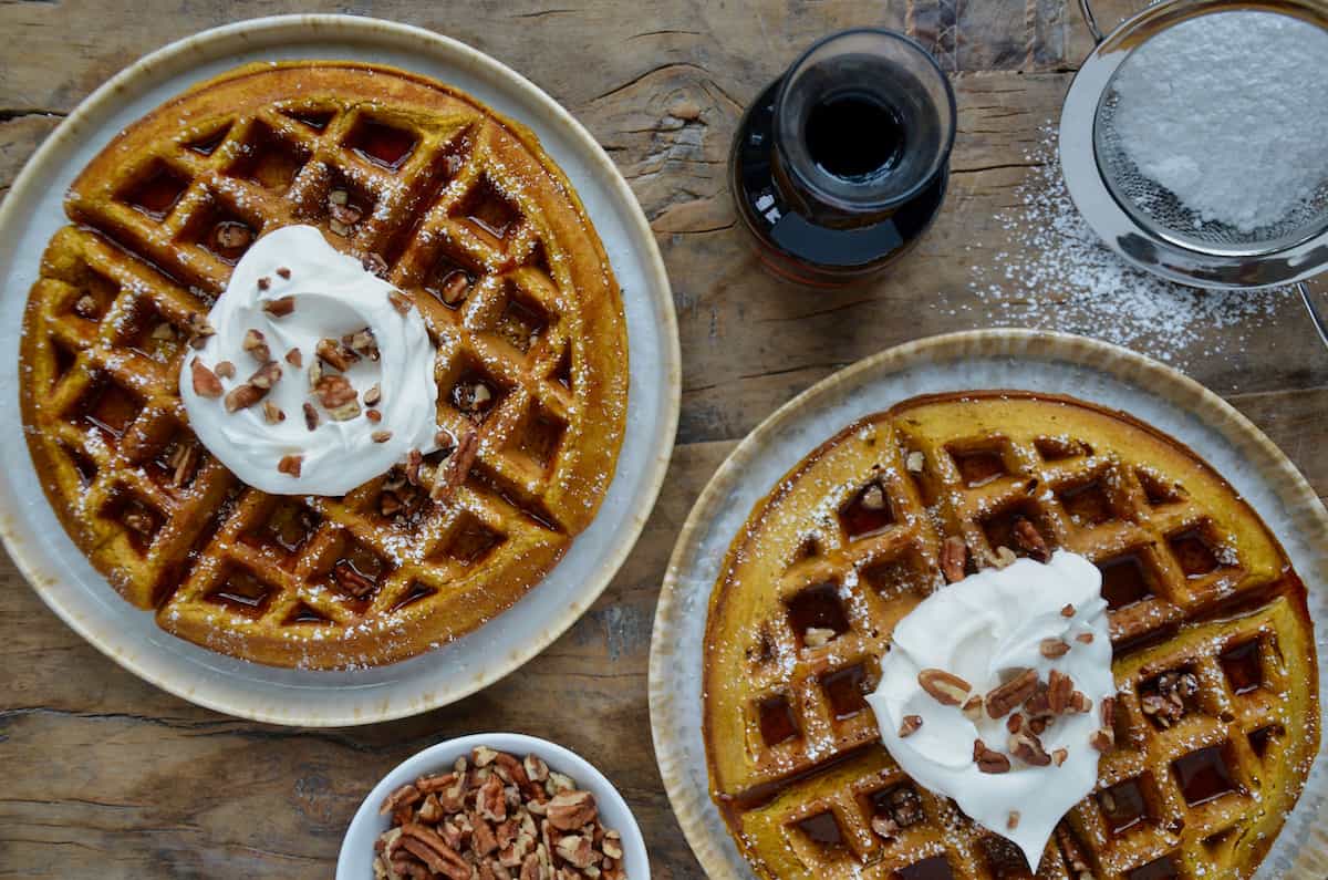 Two plates each hold a pumpkin spice waffle, topped with a dollop of whipped cream, a drizzle of syrup, and a sprinkle of pecans. Nearby is a glass pitcher of maple syrup, a dish of chopped nuts, and a sifter with powdered sugar.