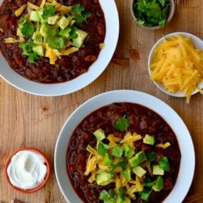 Two white bowls filled with chili and smaller bowls surrounding them containing chili toppings