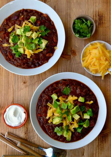 Two white bowls filled with chili and smaller bowls surrounding them containing chili toppings