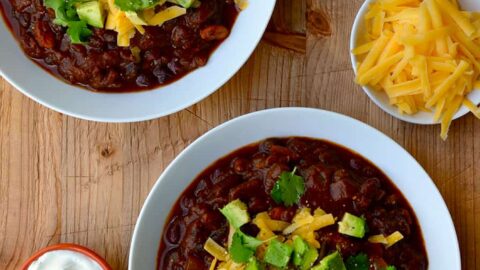 Two white bowls filled with chili and smaller bowls surrounding them containing chili toppings