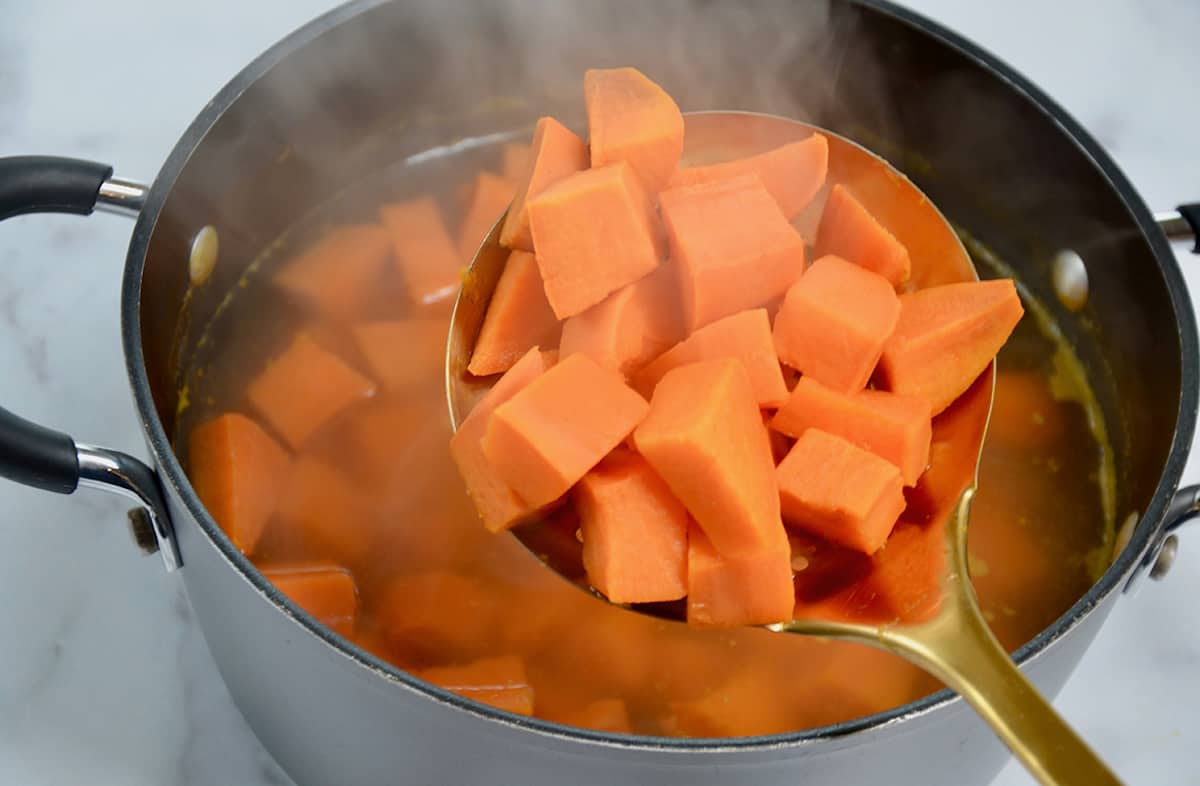 Boiled sweet potato chunks on a spoon above a dutch oven containing water and cooked sweet potatoes.