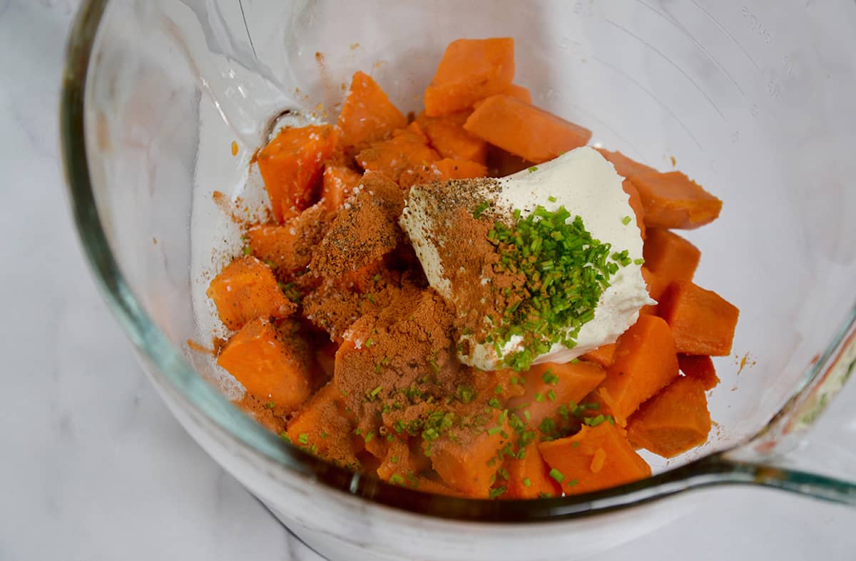 A clear bowl of a stand mixer containing boiled sweet potato chunks, cream cheese, chives and ground cinnamon.