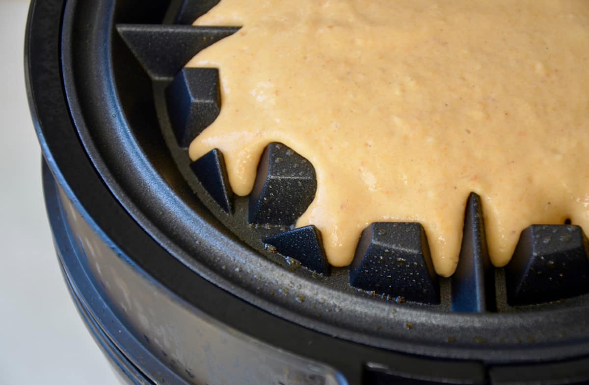 Pumpkin waffle batter is spread onto the cooking surface of a greased waffle baker.