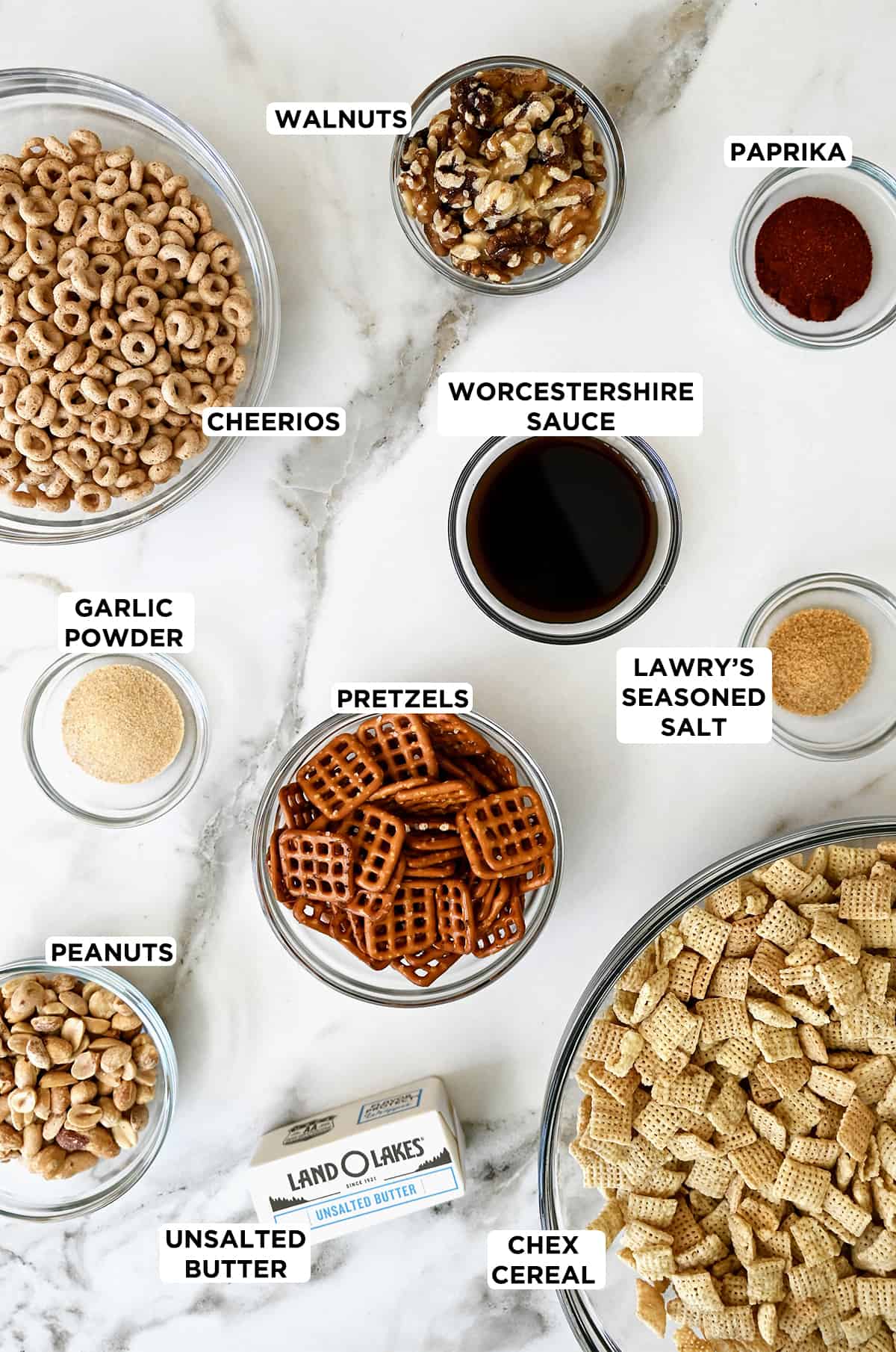 A stick of butter next to various sizes of clear bowls containing Cheerios, walnuts, paprika, Lawry’s Seasoned Salt, Chex cereal, pretzels, peanuts and garlic powder.
