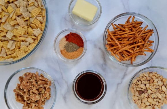 A variety of different sized bowls containing Chex Mix cereal, butter, spices, pretzels, walnuts, peanuts and Worcestershire sauce
