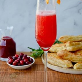 Cranberry Champagne Cocktail in Champagne flute with orange twist and fresh cranberries and pastries in the background