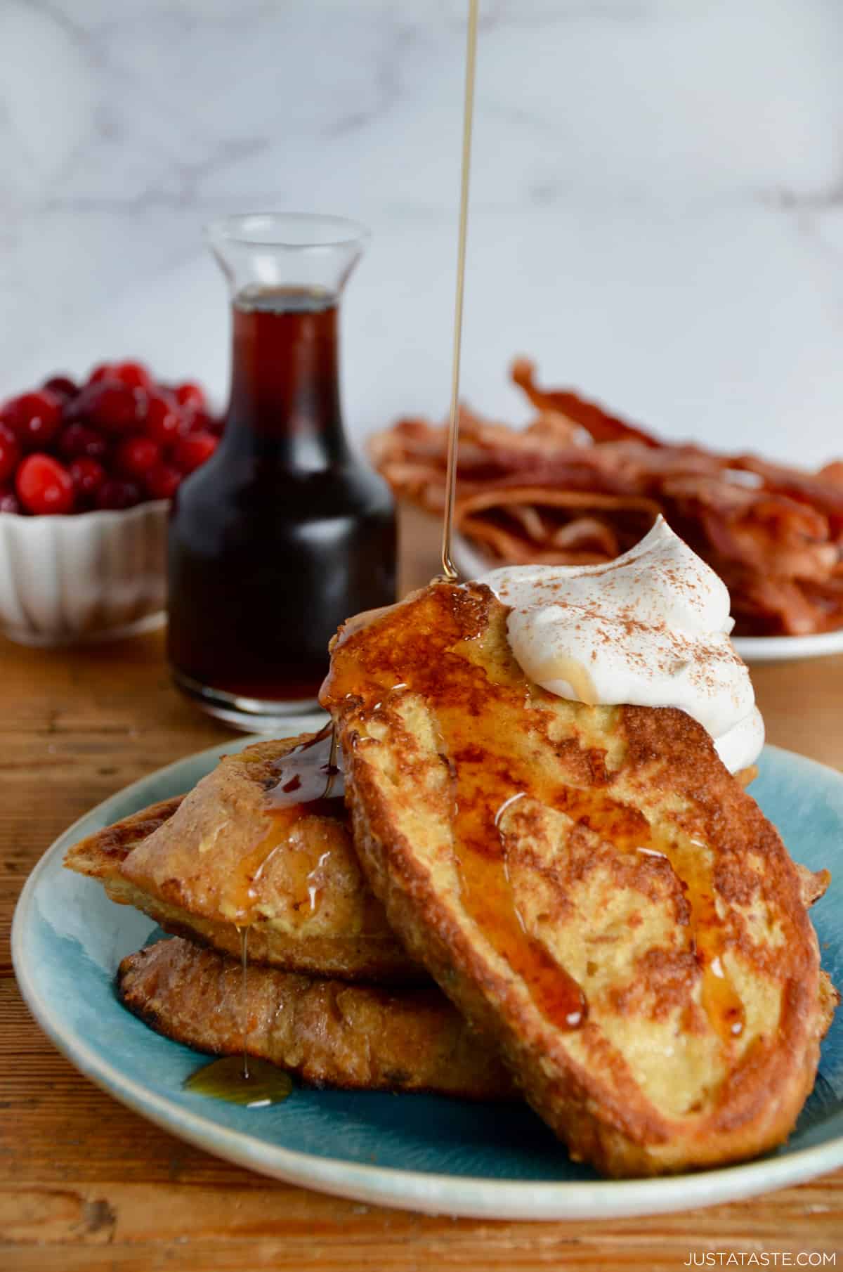 Three slices of eggnog French toast topped with whipped cream and cinnamon being drizzled with maple syrup.