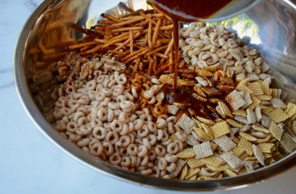 Sauce being poured over Cheerios, pretzels. walnuts and peanuts in a silver bowl