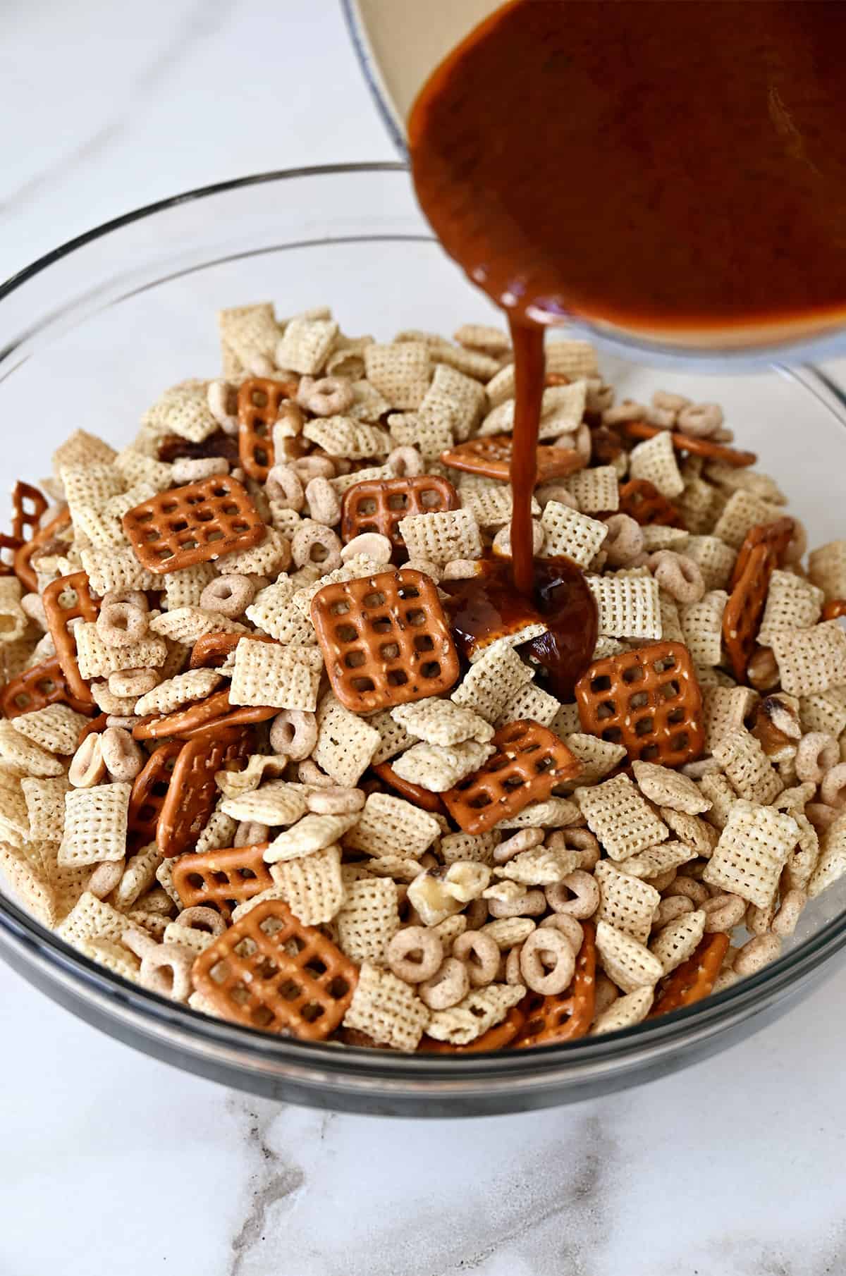 A seasoned butter mixture is being poured from a small saucepan atop Chex mix in a clear bowl.