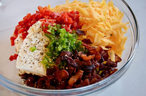 A glass bowl containing cream cheese, bacon, cheddar cheese, scallions and pimiento peppers