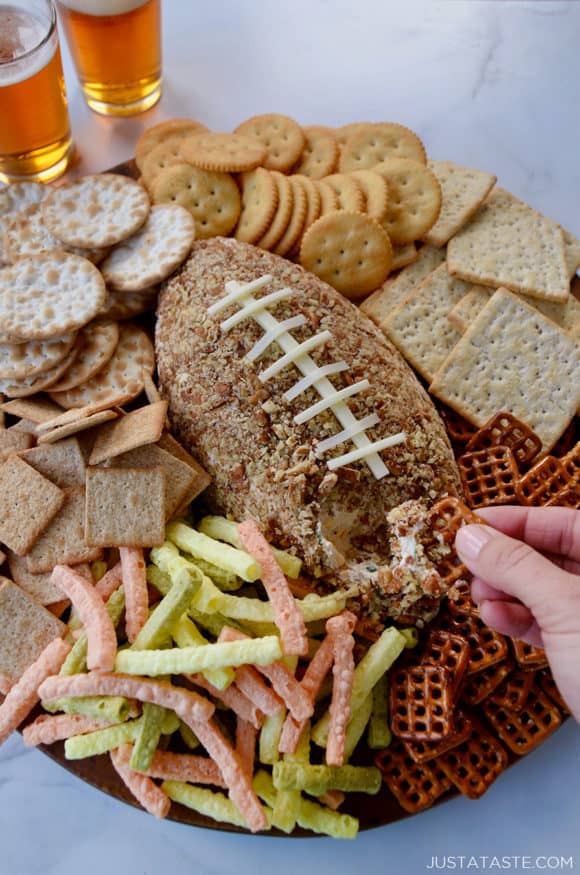 A pretzel being dipped into a cheese ball with snacks surrounding it