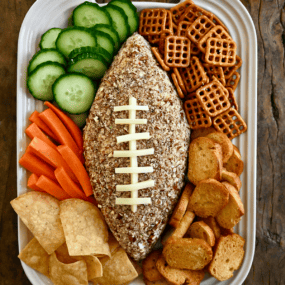 A football-shaped cheese ball on a white platter surrounded by cucumber slices, pretzels, pita chips, tortilla chips and carrot sticks.