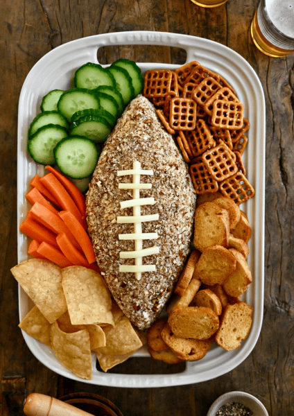 A football-shaped cheese ball on a white platter surrounded by cucumber slices, pretzels, pita chips, tortilla chips and carrot sticks.