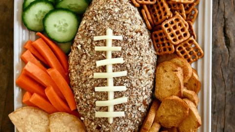 A football-shaped cheese ball on a white platter surrounded by cucumber slices, pretzels, pita chips, tortilla chips and carrot sticks.