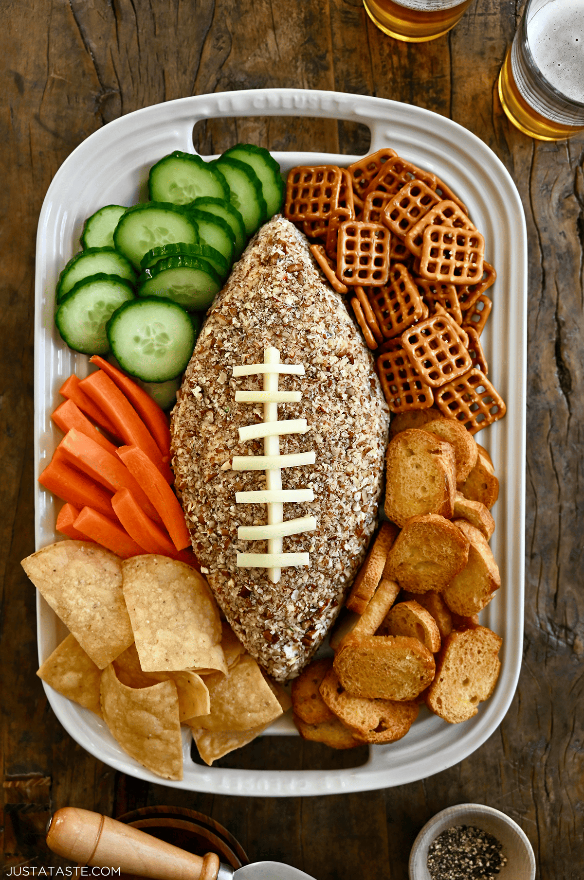 A football-shaped cheese ball on a white platter surrounded by cucumber slices, pretzels, pita chips, tortilla chips and carrot sticks.