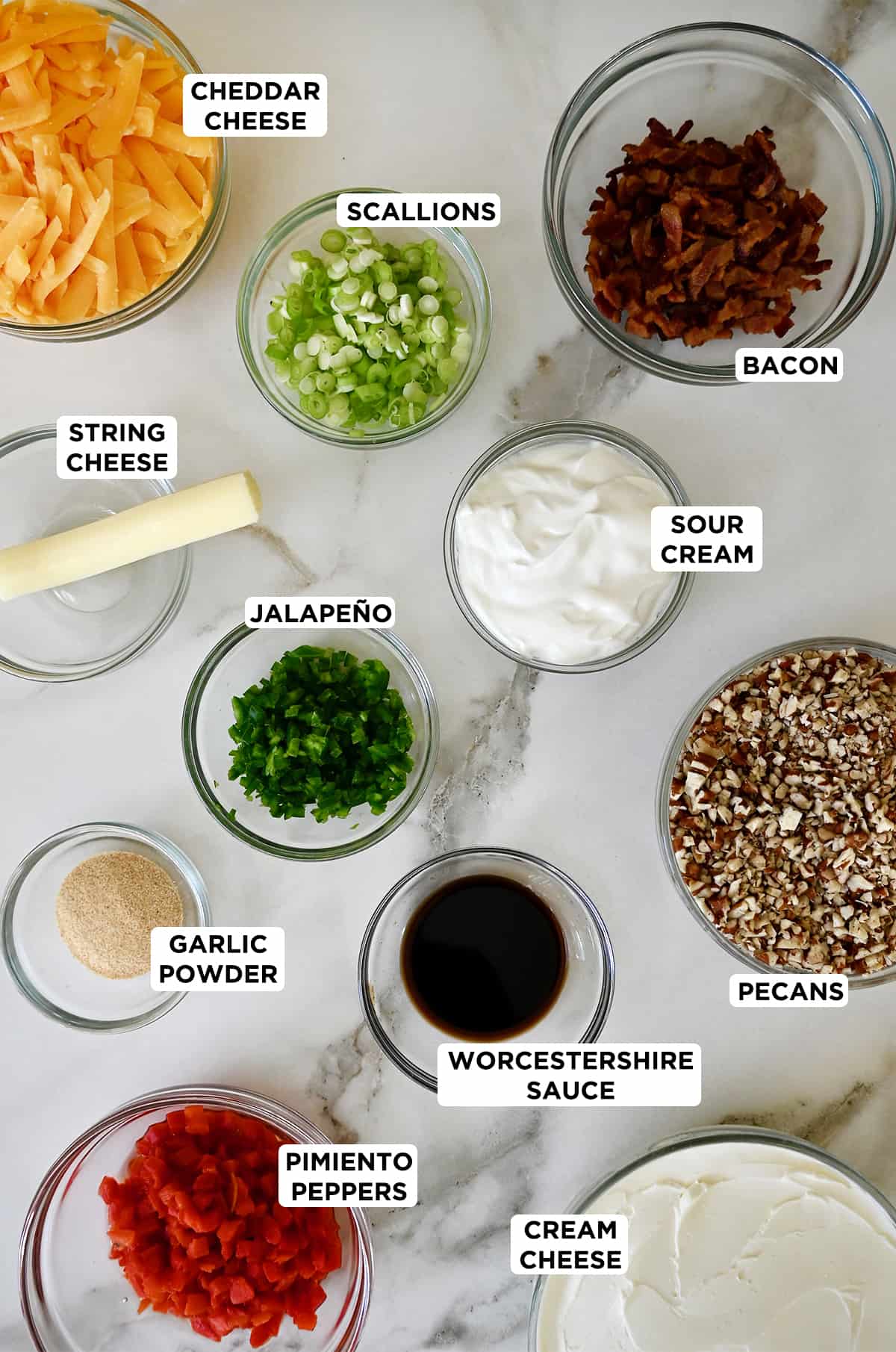Various sizes of glass bowls containing ingredients to make a cheese ball, including shredded cheddar cheese, sliced scallions, crispy bacon, sour cream, diced jalapeño, string cheese, garlic powder, Worcestershire sauce, crushed pecans and pimento peppers.