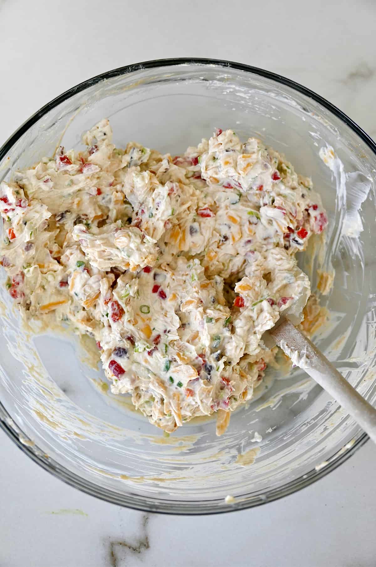 A glass bowl containing a cheddar, cream cheese and bacon mixture with a spatula.