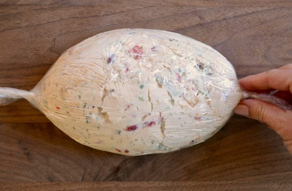 A wood cutting board with a cheese ball wrapped in plastic wrap