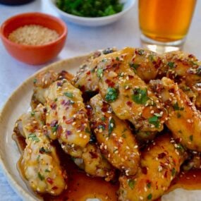 A white plate containing chicken wings with sesame seeds, cilantro and a beer in the background