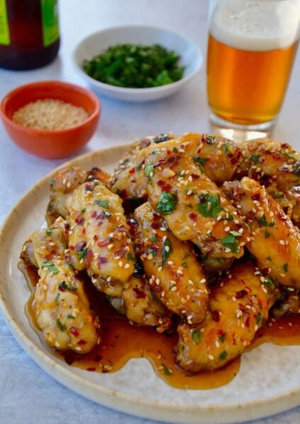A white plate containing chicken wings with sesame seeds, cilantro and a beer in the background