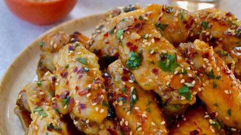 A white plate containing chicken wings with sesame seeds, cilantro and a beer in the background