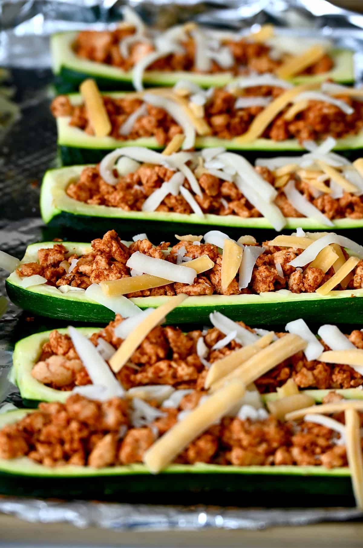 Stuffed zucchini boats topped with shredded cheese on a foil-lined baking sheet before melting the cheese.