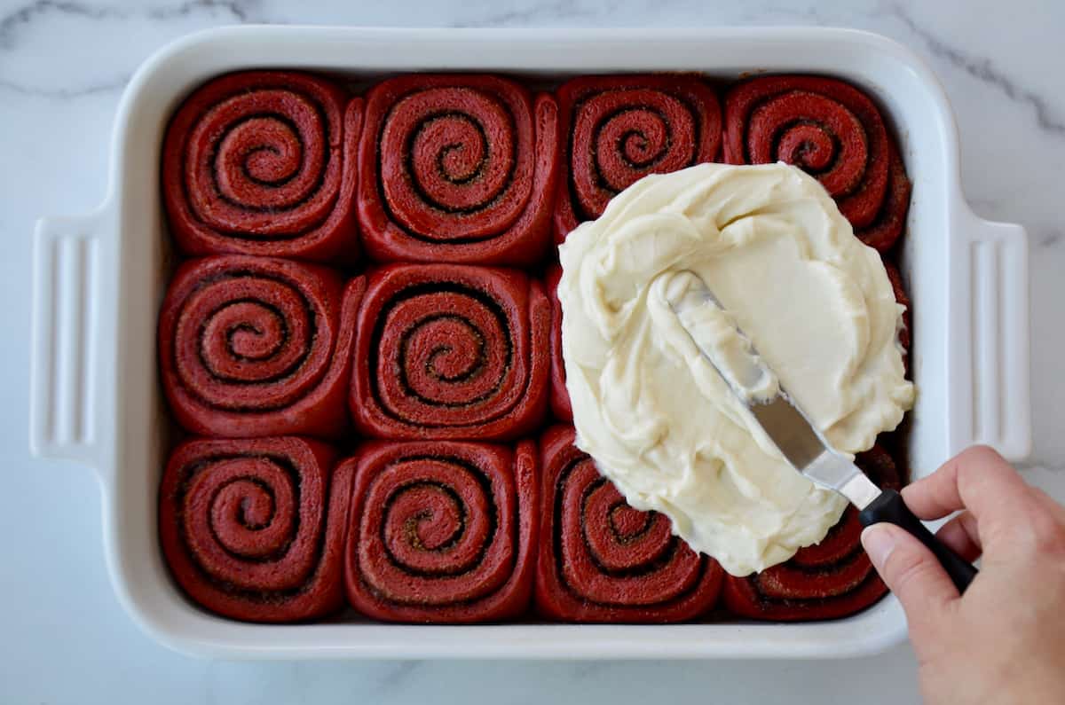 A hand holding an offset spatula slathers cream cheese frosting atop red velvet cinnamon rolls in a white baking dish.