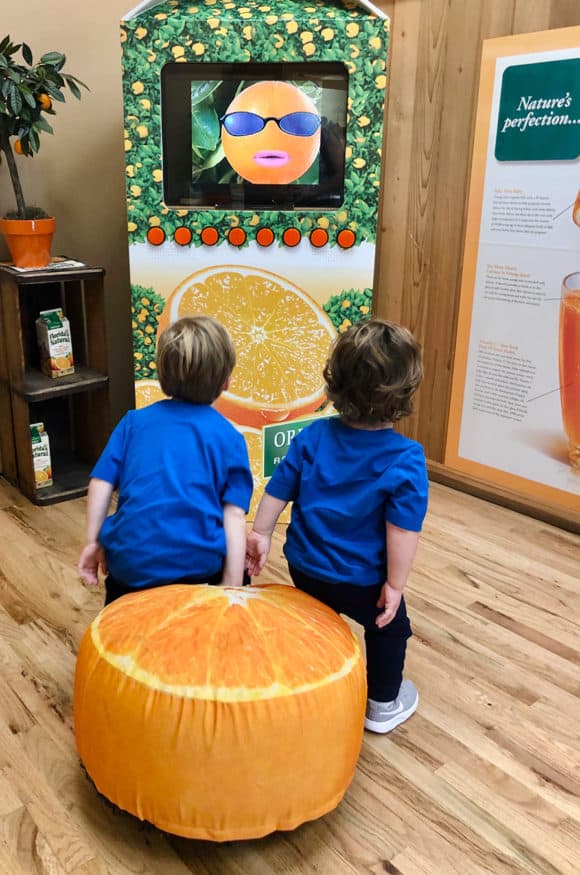 Two toddler boys sitting on an orange pillow watching a TV screen