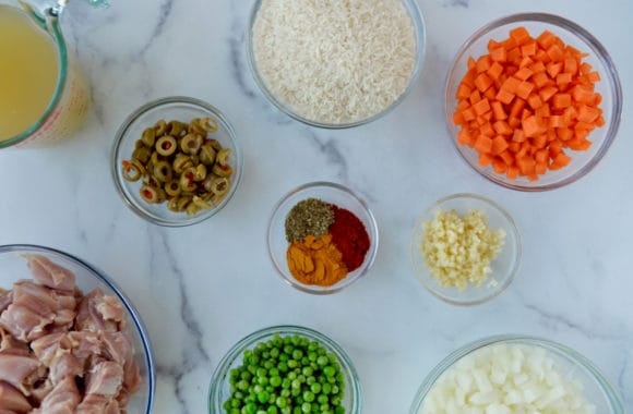 Various sized bowls containing chicken stock, green olives, diced onions and carrots, minced garlic, spices, frozen peas, basmati rice and uncooked cubed chicken thighs 