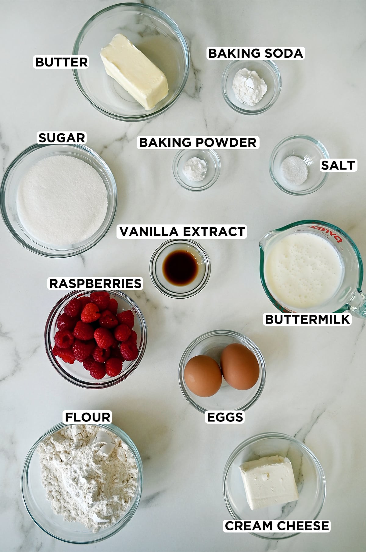 Various sizes of glass bowls containing ingredients needed to make a coffee cake, including a stick of butter, baking soda and baking powder, salt, sugar, vanilla extract, buttermilk, two eggs, flour, cream cheese, and fresh raspberries.
