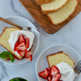 Cream Cheese Pound Cake sliced on cutting board with two slices on dessert plates with whipped cream and fresh strawberries