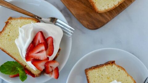 Cream Cheese Pound Cake sliced on cutting board with two slices on dessert plates with whipped cream and fresh strawberries