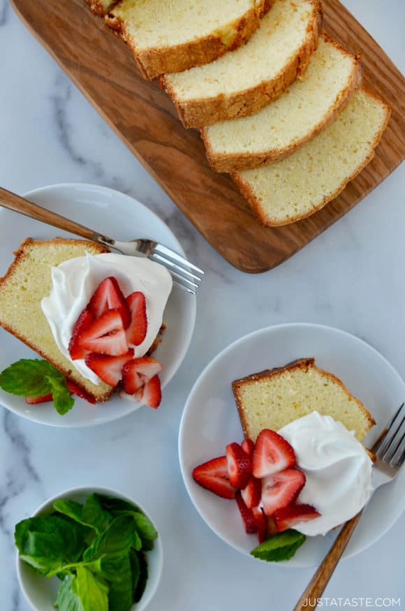 Cream Cheese Pound Cake sliced on cutting board with two slices on dessert plates with whipped cream and fresh strawberries