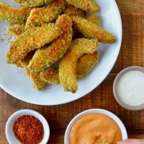 Hand dipping avocado fry into aioli sauce next to plate containing Crispy Baked Avocado Fries and small ramekins with spices and ranch