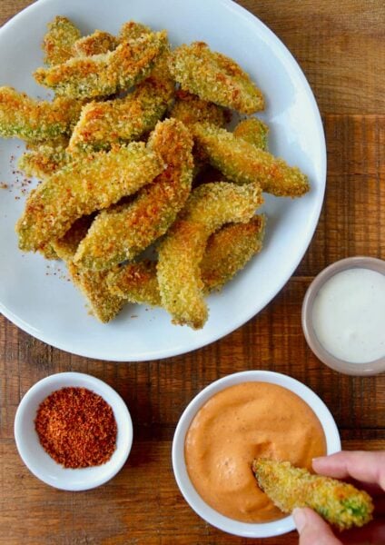 Hand dipping avocado fry into aioli sauce next to plate containing Crispy Baked Avocado Fries and small ramekins with spices and ranch