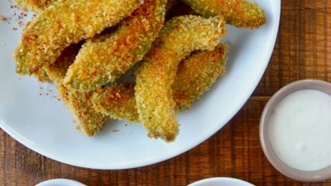 Hand dipping avocado fry into aioli sauce next to plate containing Crispy Baked Avocado Fries and small ramekins with spices and ranch