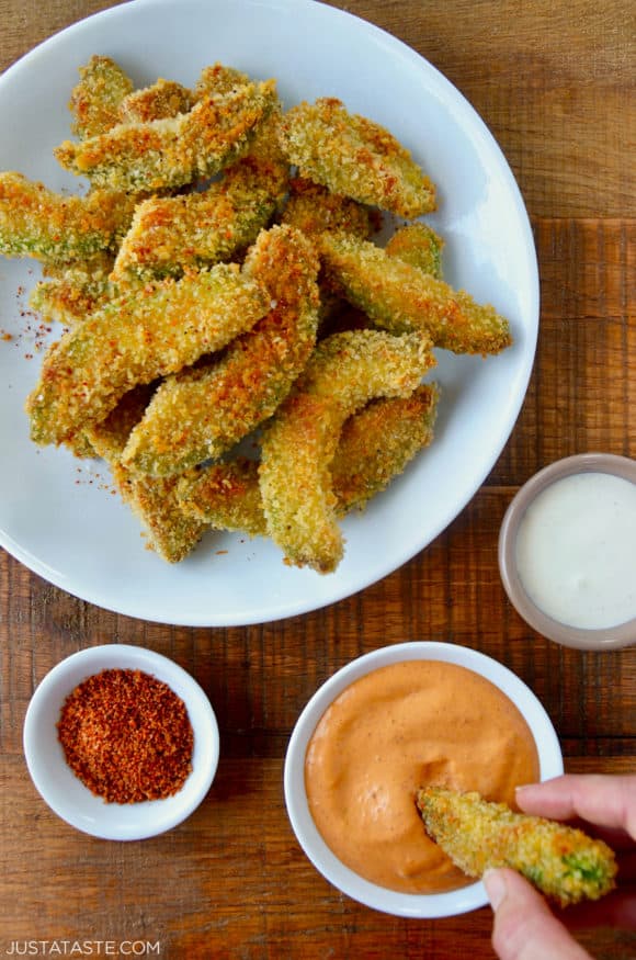 Hand dipping avocado fry into aioli sauce next to plate containing Crispy Baked Avocado Fries and small ramekins with spices and ranch