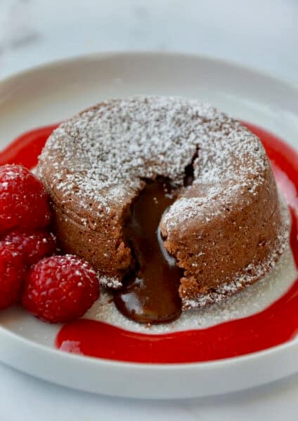 A chocolate lava cake with a molten chocolate center oozing out onto a dessert plate. The lava cake is topped with powdered sugar and garnished with fresh raspberries and a raspberry sauce.