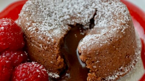 A chocolate lava cake with a molten chocolate center oozing out onto a dessert plate. The lava cake is topped with powdered sugar and garnished with fresh raspberries and a raspberry sauce.