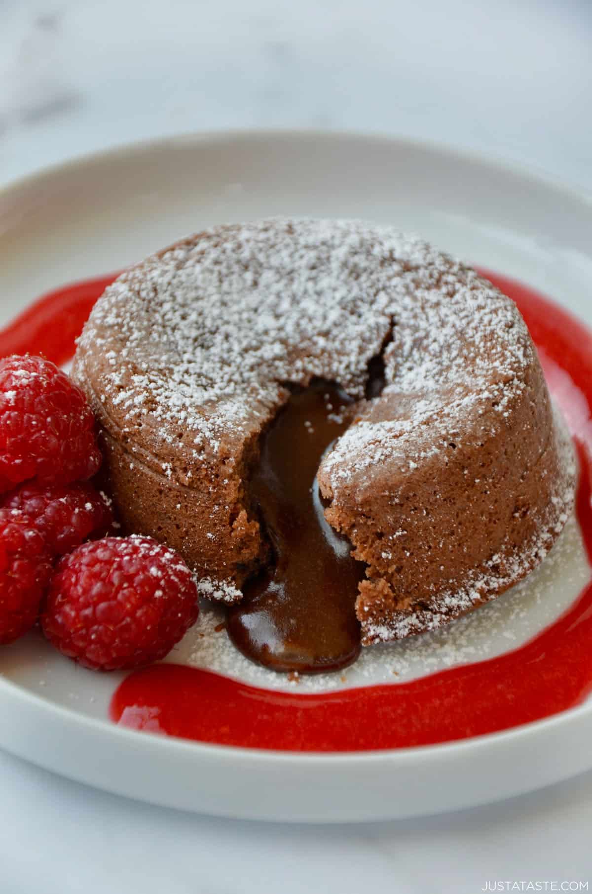 A chocolate lava cake with a molten chocolate center oozing out onto a dessert plate. The lava cake is topped with powdered sugar and garnished with fresh raspberries and a raspberry sauce.