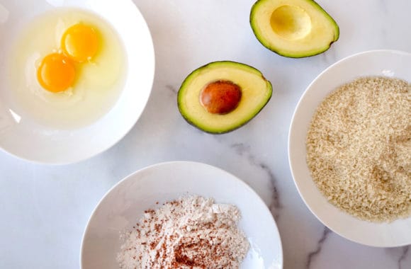 Bowls containing cracked eggs, panko breadcrumbs and flour with spices next to an avocado cut open in half