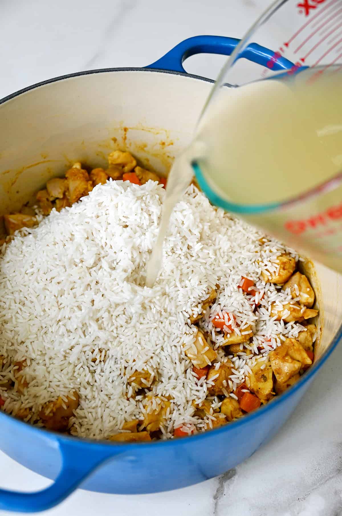 Chicken stock being poured over long-grain white rice in a Dutch oven.