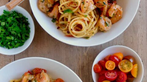 Two bowls with Quick Shrimp Scampi next to a glass of wine and small bowls containing cherry tomatoes, chili flakes and parsley