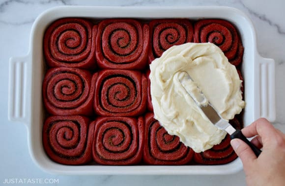 A white baking dish with red velvet cinnamon rolls and a spatula slathering cream cheese frosting on top